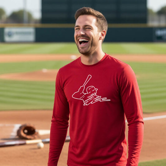 A man smiling in front of a baseball diamond wearing a red long-sleeve t-shirt, with a white graphic of an outline of a baseball player up to bat, printed on the front.