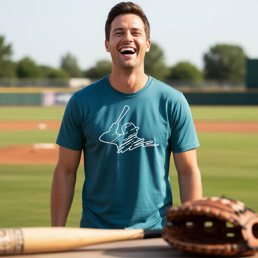 A man smiling in front of a baseball diamond wearing a teal short-sleeve t-shirt, with a white graphic of an outline of a baseball player up to bat, printed on the front.
