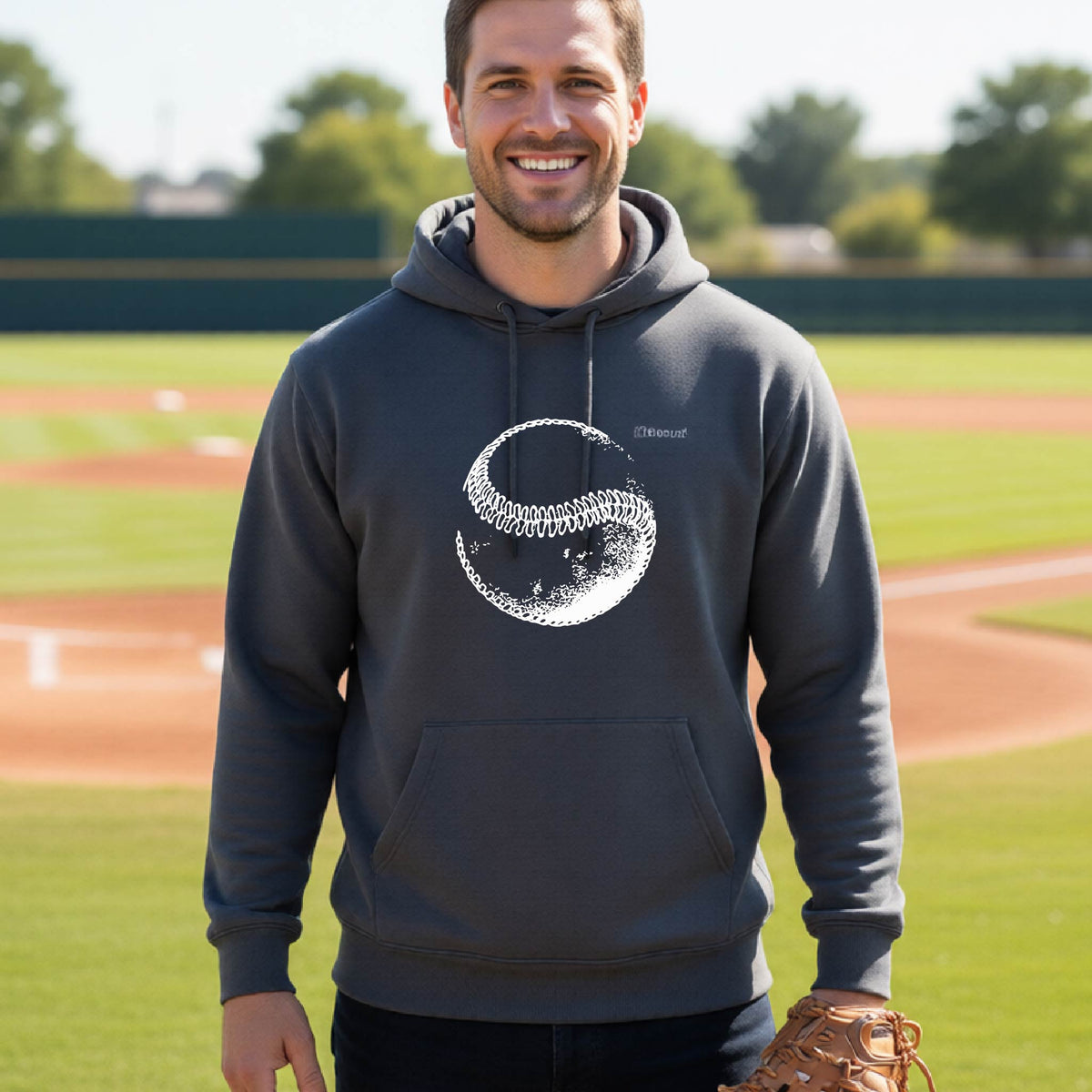A man smiling in front of a baseball diamond wearing a dark gray hoodie pullover, with a white artistic graphic of a baseball, printed on the front.