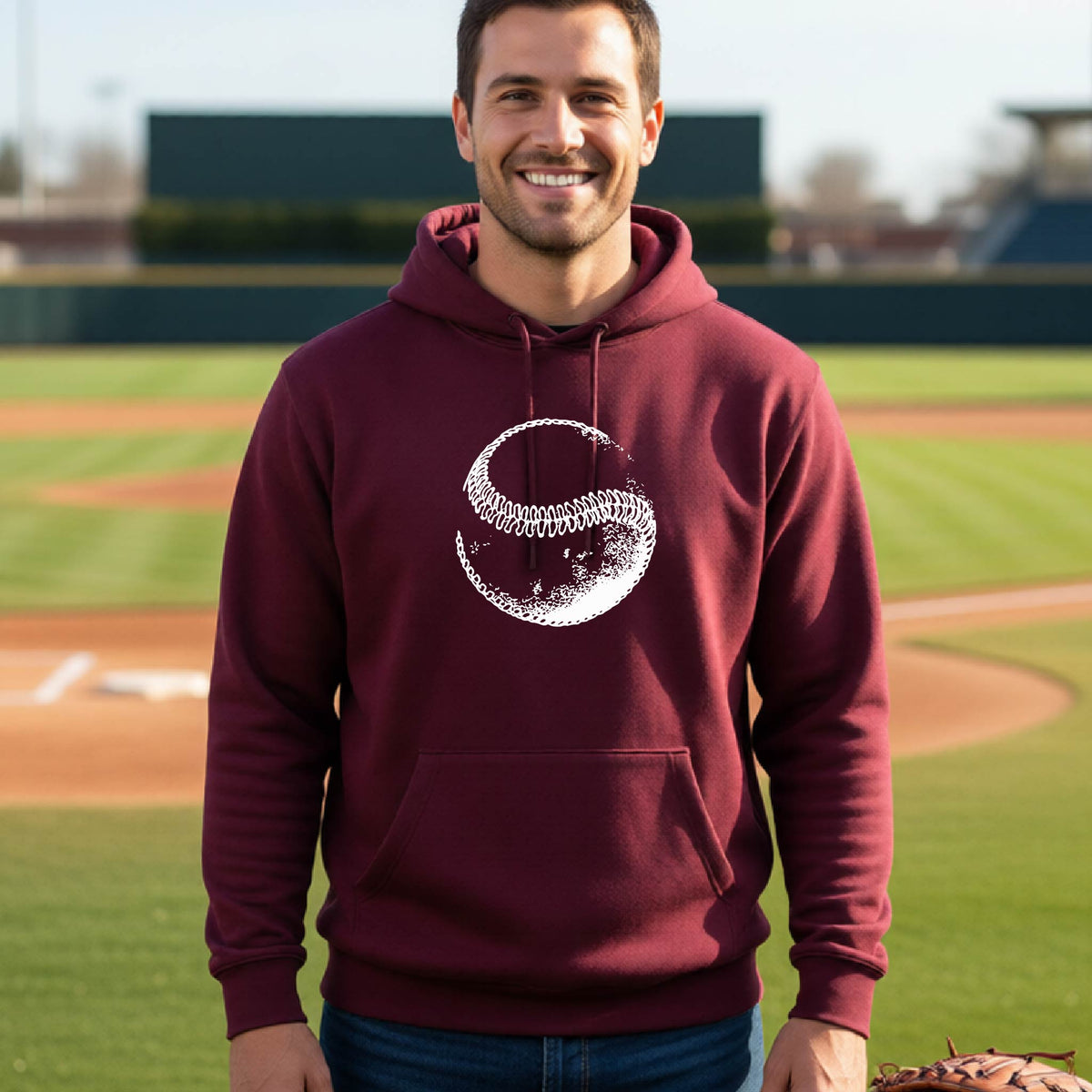 A man smiling in front of a baseball diamond wearing a maroon hoodie pullover, with a white artistic graphic of a baseball, printed on the front.