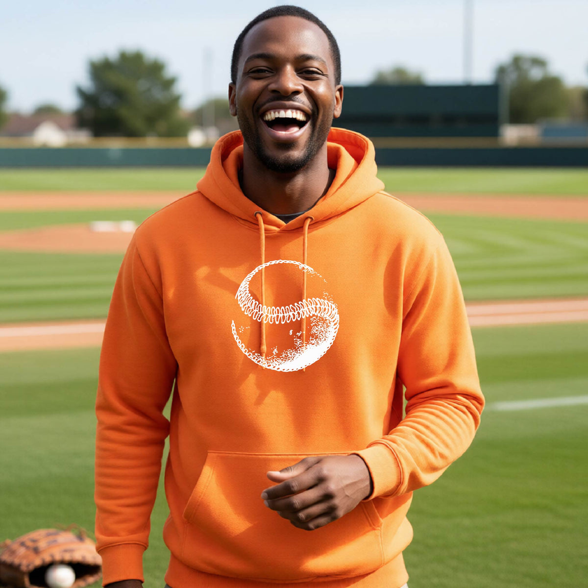 A man smiling in front of a baseball diamond wearing an orange hoodie pullover, with a white artistic graphic of a baseball, printed on the front.