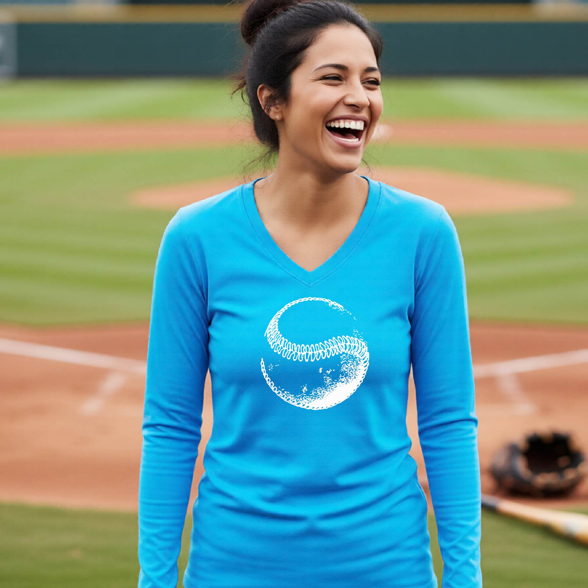 A woman smiling in front of a baseball diamond wearing a bright blue long-sleeve t-shirt, with a white artistic graphic of a baseball printed on the front.