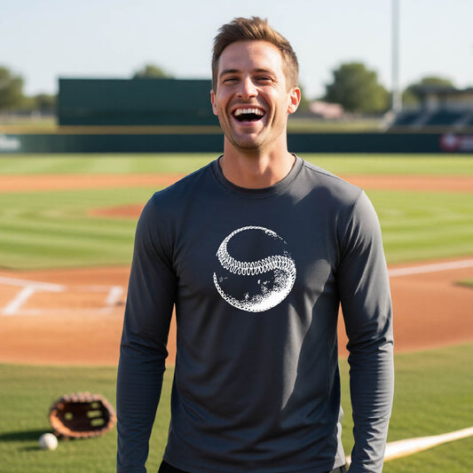 A man smiling in front of a baseball diamond wearing a dark gray long-sleeve t-shirt, with a white artistic graphic of a baseball printed on the front.