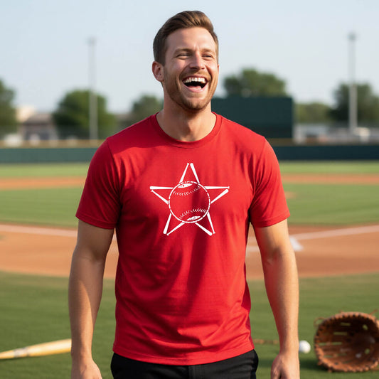 A man smiling in front of a baseball diamond wearing a red short-sleeve t-shirt, with a white graphic of a star made of baseball bats and a ball in the middle, printed on the front.
