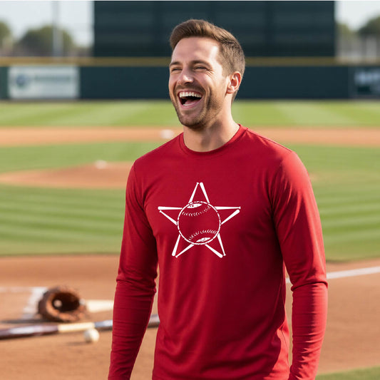 A man smiling in front of a baseball diamond wearing a red long-sleeve t-shirt, with a white graphic of a star made of baseball bats and a ball in the middle, printed on the front.