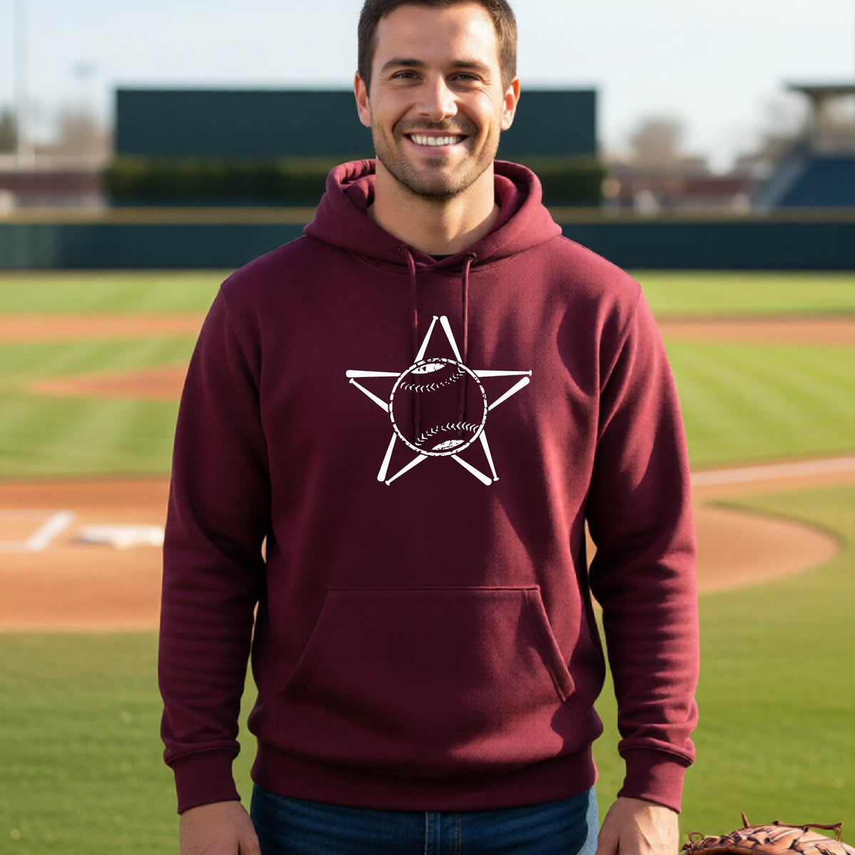 A man smiling in front of a baseball diamond wearing a maroon hoodie pullover, with a white graphic of a star made of baseball bats and a ball in the middle, printed on the front.