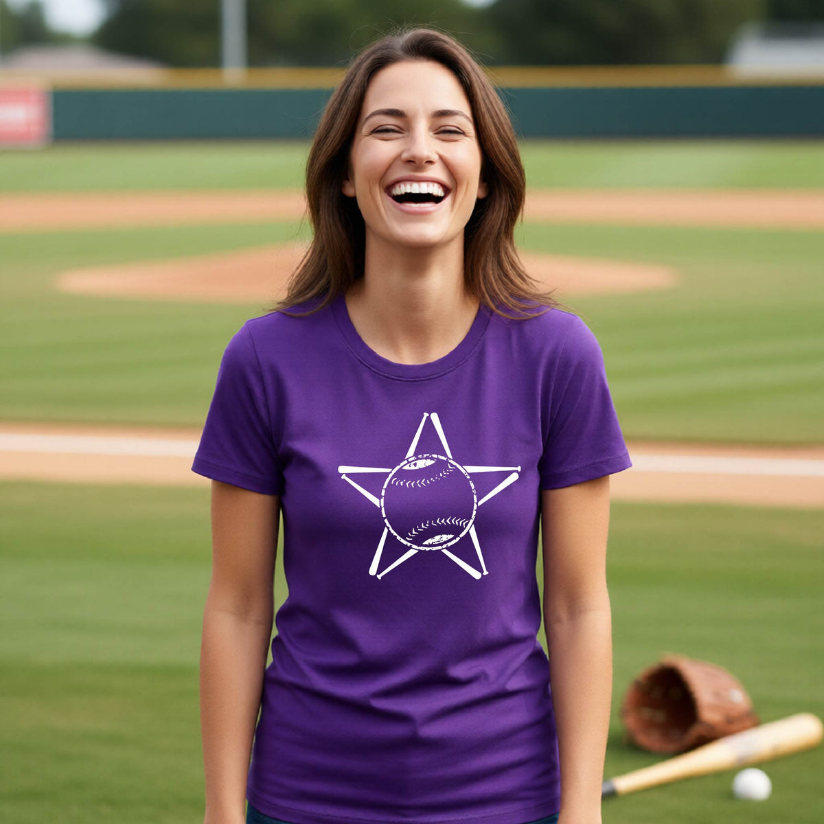 A woman smiling in front of a baseball diamond wearing a purple short-sleeve t-shirt, with a white graphic of a star made of baseball bats and a ball in the middle, printed on the front.