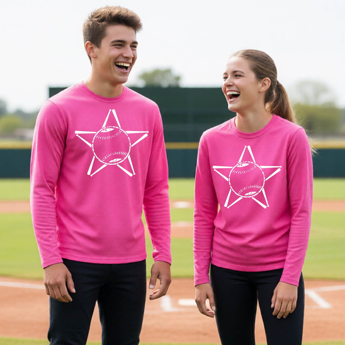 A man smiling in front of a baseball diamond wearing a pink long-sleeve t-shirt, with a white graphic of a star made of baseball bats and a ball in the middle, printed on the front.