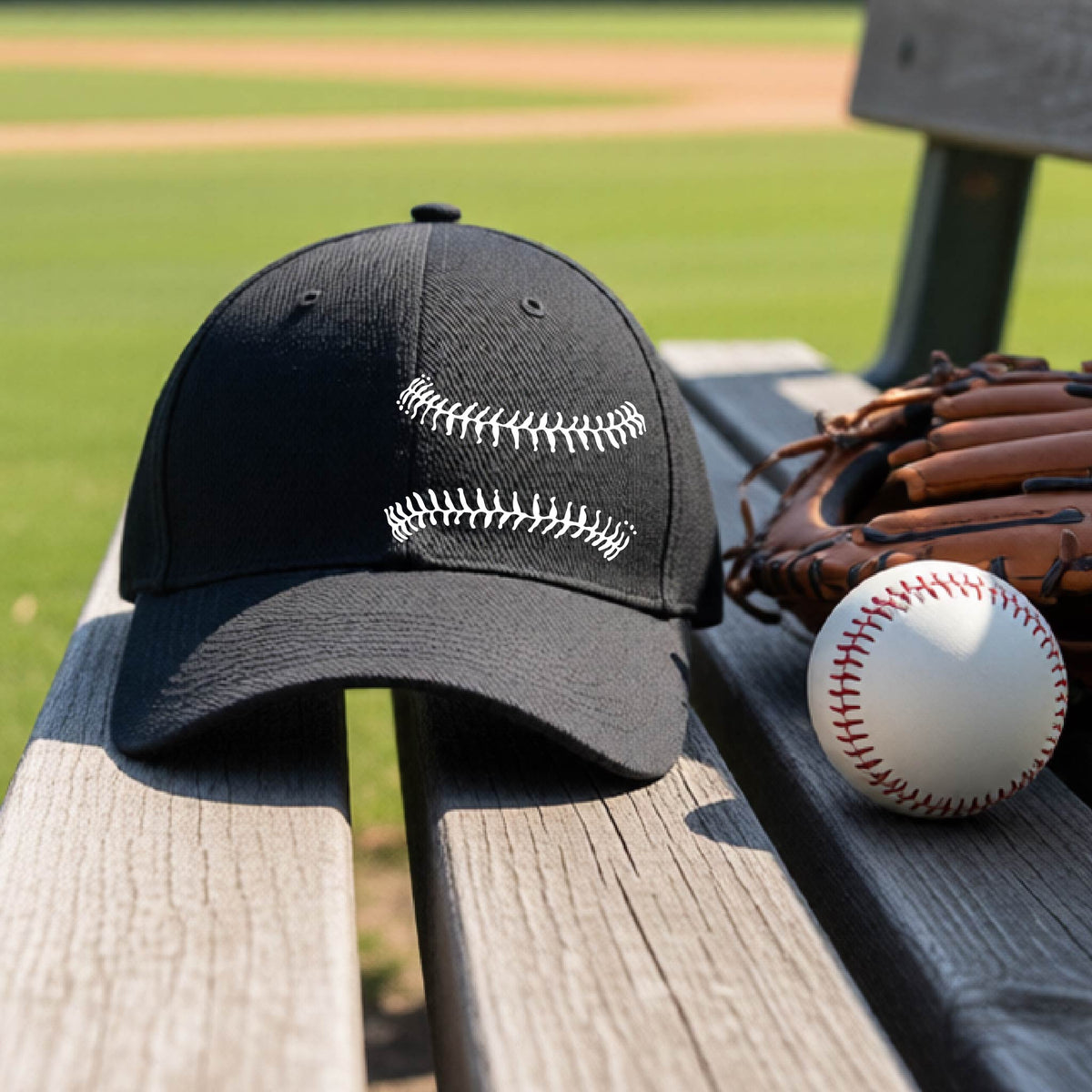 A closeup of a black baseball hat on an outdoor bench in front of a baseball diamond, with a white graphic of baseball stitches, printed on the front.