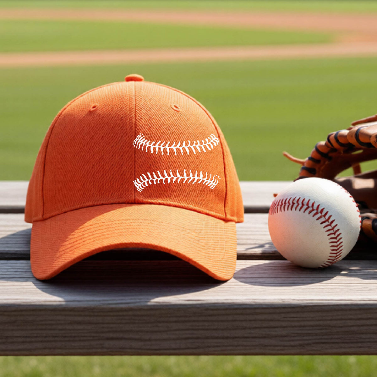 A closeup of an orange baseball hat on an outdoor bench in front of a baseball diamond, with a white graphic of baseball stitches, printed on the front.