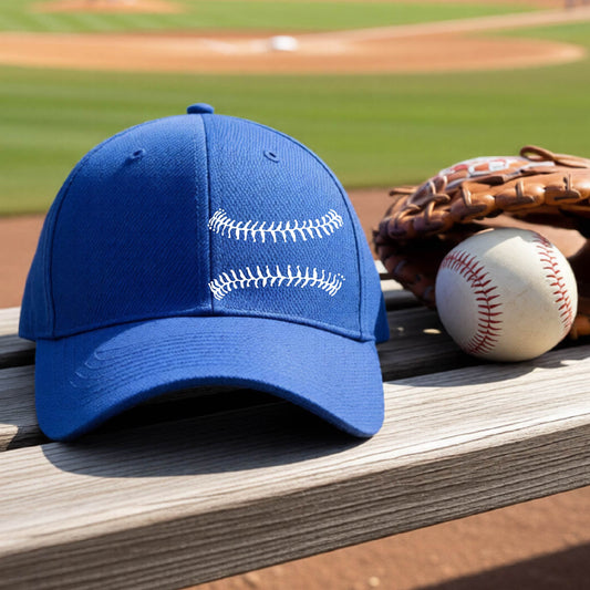 A closeup of a royal blue baseball hat on an outdoor bench in front of a baseball diamond, with a white graphic of baseball stitches, printed on the front.