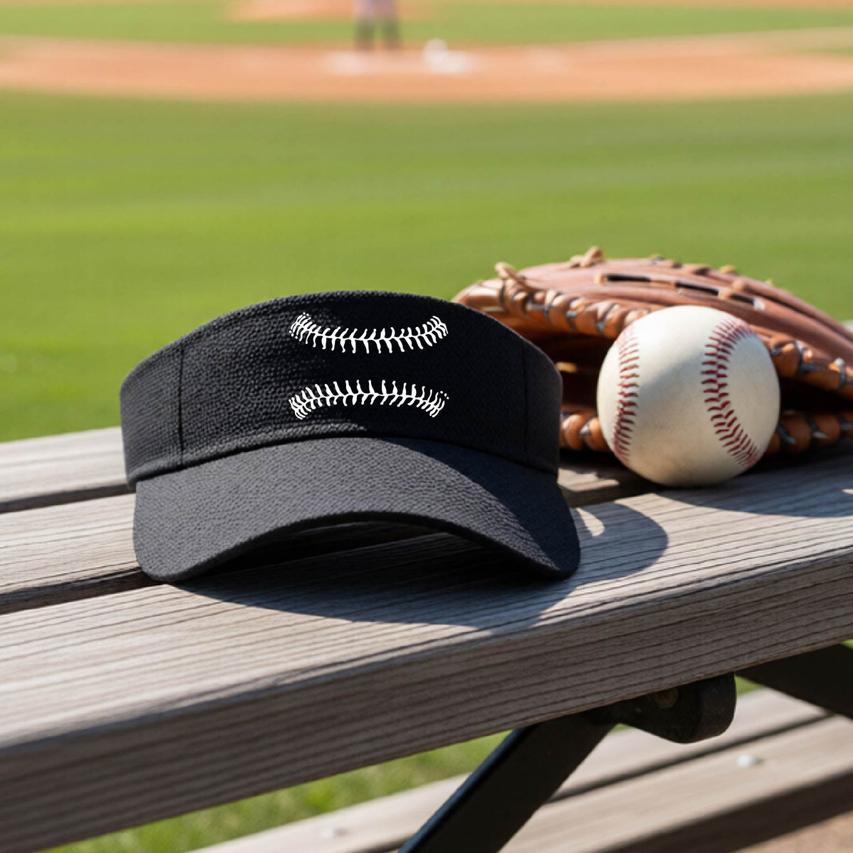 A closeup of a black baseball visor on an outdoor bench in front of a baseball diamond, with a white graphic of baseball stitches, printed on the front.