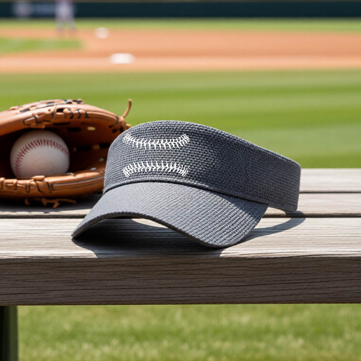 A closeup of a graphite gray baseball visor on an outdoor bench in front of a baseball diamond, with a white graphic of baseball stitches, printed on the front.
