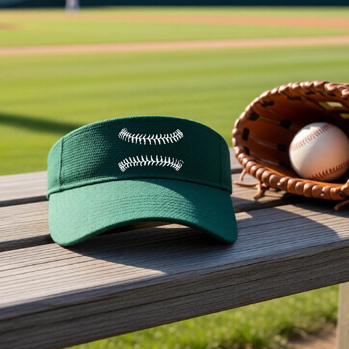 A closeup of a green baseball visor on an outdoor bench in front of a baseball diamond, with a white graphic of baseball stitches, printed on the front.