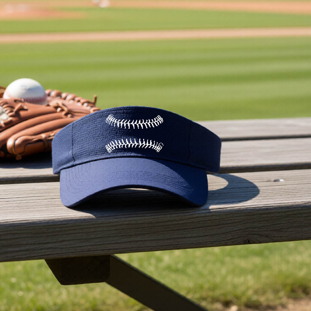 A closeup of a navy blue baseball visor on an outdoor bench in front of a baseball diamond, with a white graphic of baseball stitches, printed on the front.