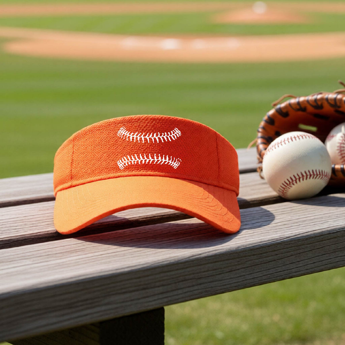A closeup of an orange baseball visor on an outdoor bench in front of a baseball diamond, with a white graphic of baseball stitches, printed on the front.