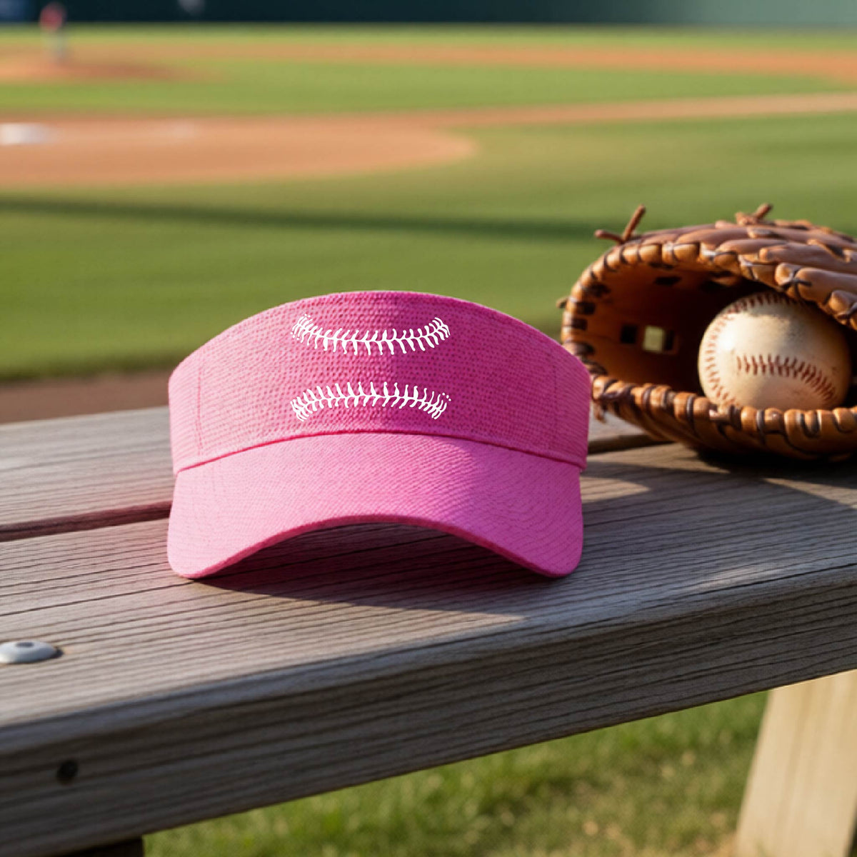 A closeup of a pink baseball visor on an outdoor bench in front of a baseball diamond, with a white graphic of baseball stitches, printed on the front.