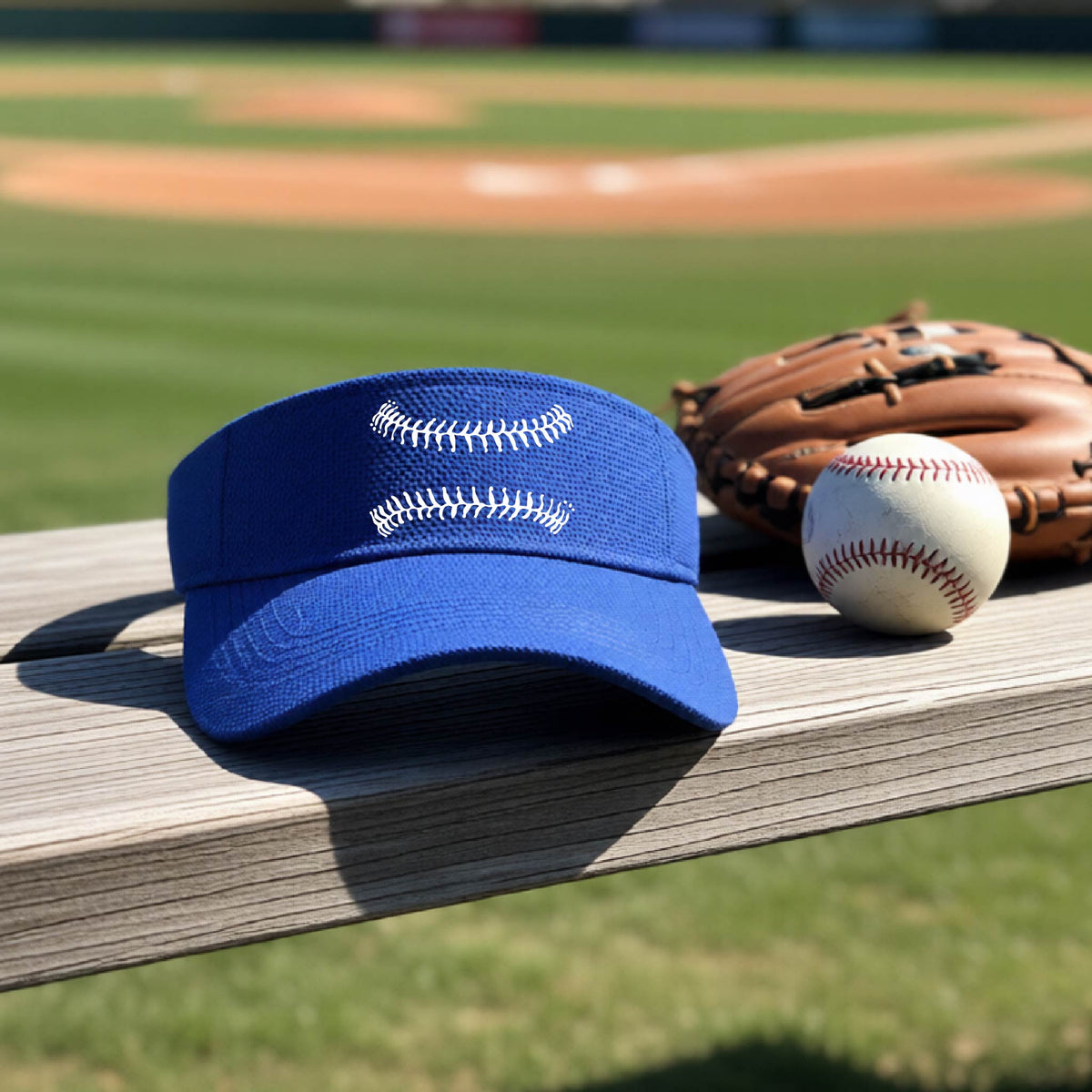 A closeup of a royal blue baseball visor on an outdoor bench in front of a baseball diamond, with a white graphic of baseball stitches, printed on the front.