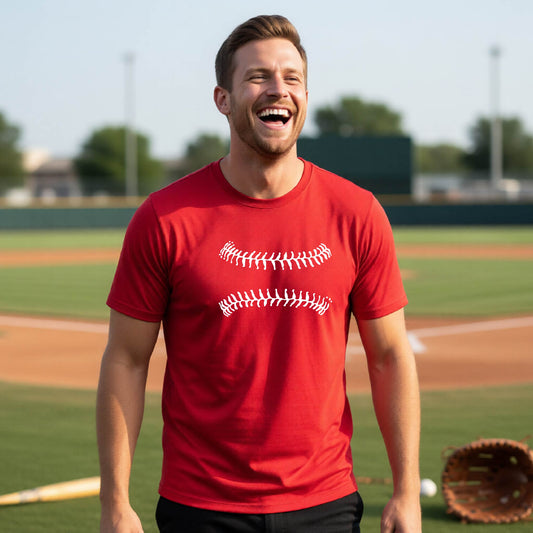 A man smiling in front of a baseball diamond wearing a red short-sleeve t-shirt, with a white graphic of baseball stitches, printed on the front.