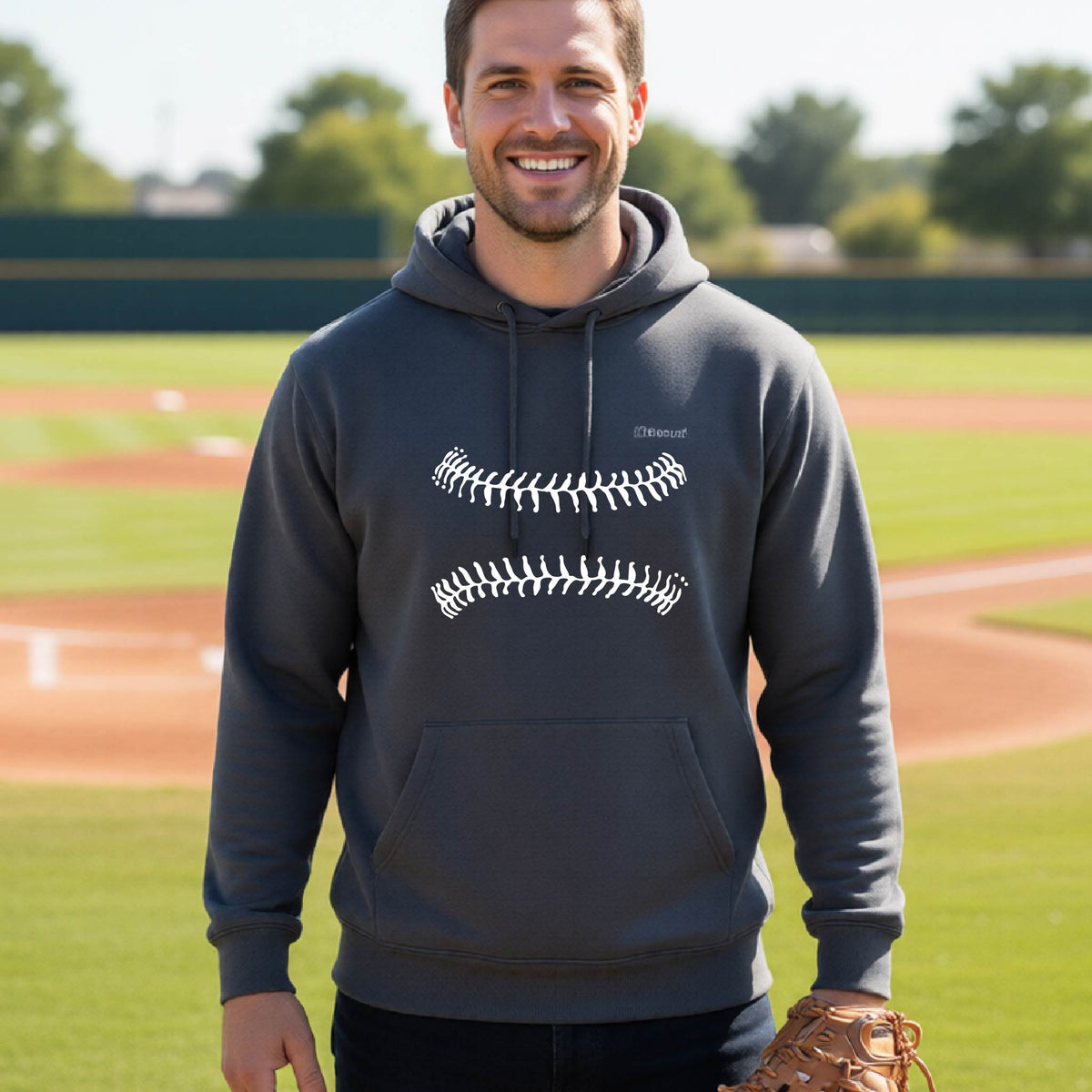A woman smiling in front of a baseball diamond wearing a dark gray hoodie pullover, with a white graphic of baseball stitches, printed on the front.