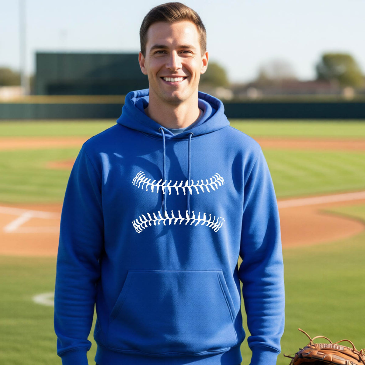 A woman smiling in front of a baseball diamond wearing a royal blue hoodie pullover, with a white graphic of baseball stitches, printed on the front.