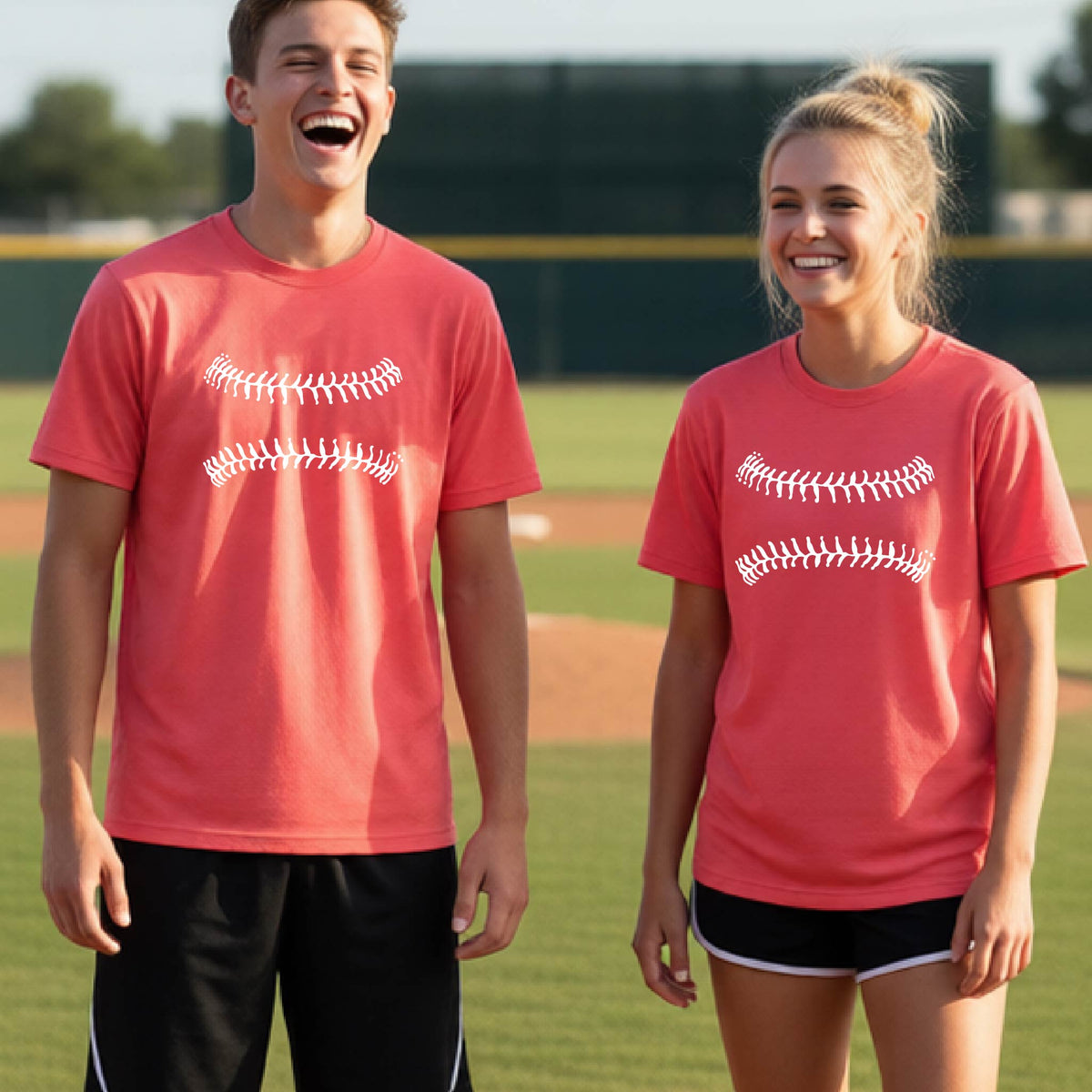 Two youth athletes wearing matching shirts, smiling in front of a baseball diamond wearing coral colored short-sleeve t-shirts with a white graphic of baseball stitches, printed on the front.