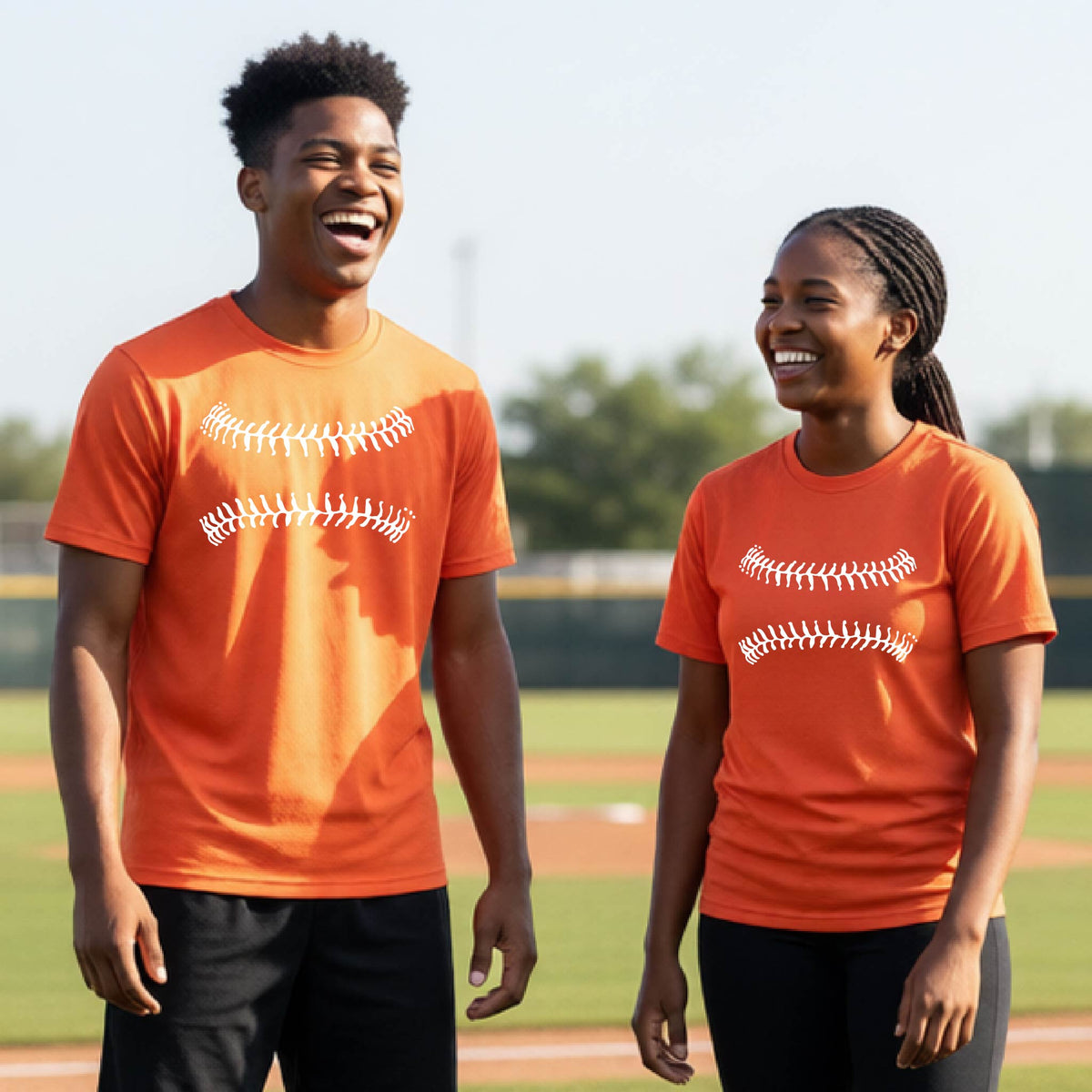 Two youth athletes wearing matching shirts, smiling in front of a baseball diamond wearing orange short-sleeve t-shirts with a white graphic of baseball stitches, printed on the front.