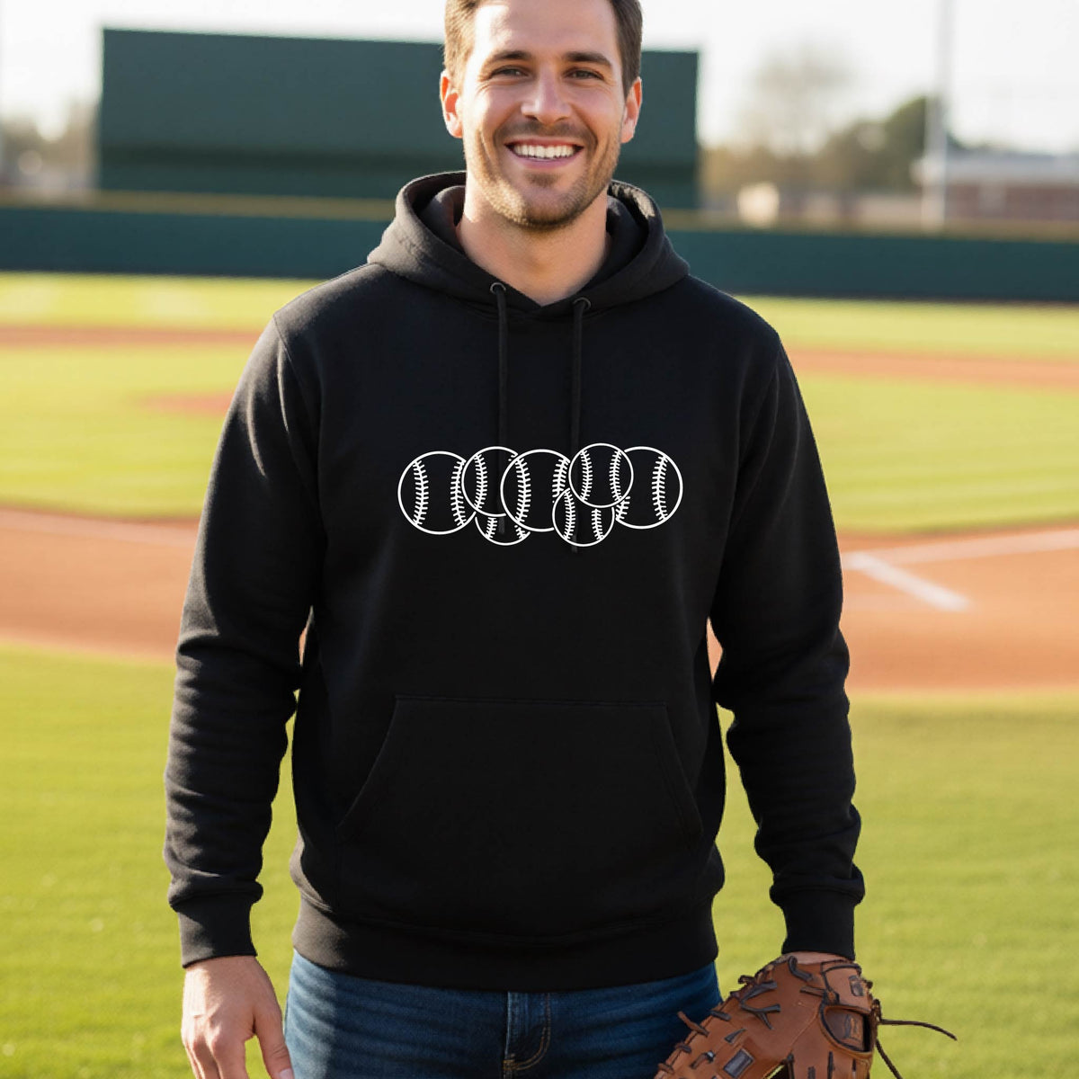 A man smiling in front of a baseball diamond wearing a black hoodie pullover, with a white graphic of 6 sketched baseballs overlapping, printed on the front of the shirt