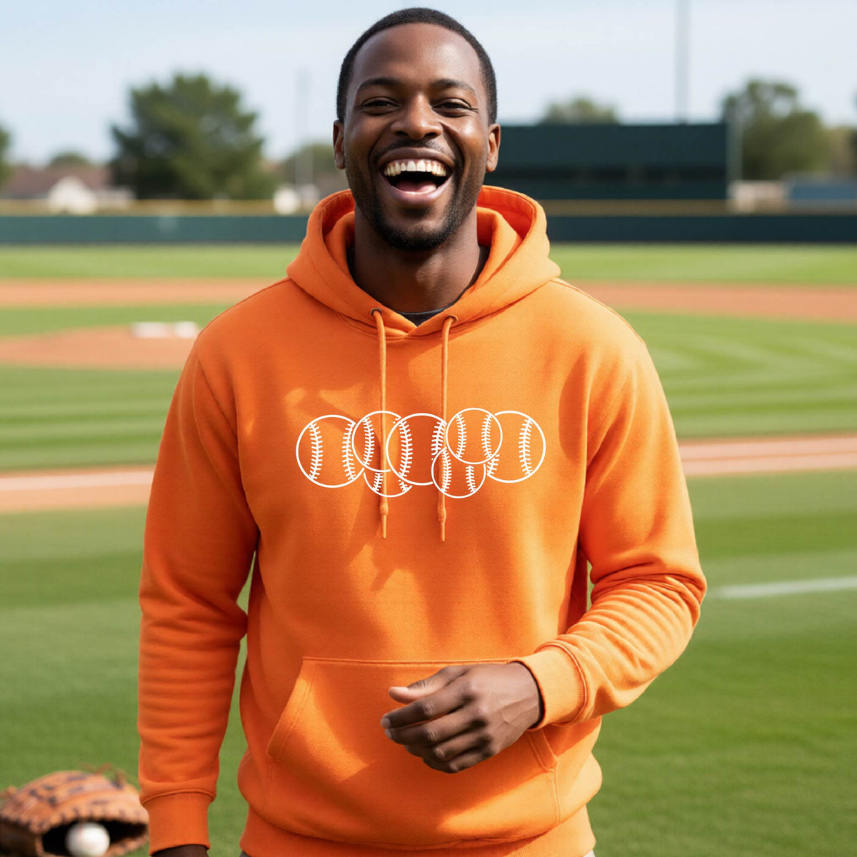 A man smiling in front of a baseball diamond wearing an orange hoodie pullover, with a white graphic of 6 sketched baseballs overlapping, printed on the front of the shirt