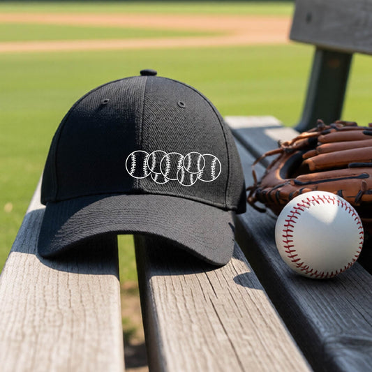 A closeup of a black baseball hat on an outdoor bench in front of a baseball diamond, with a white graphic of 6 sketched baseballs overlapping, printed on the front of the shirt