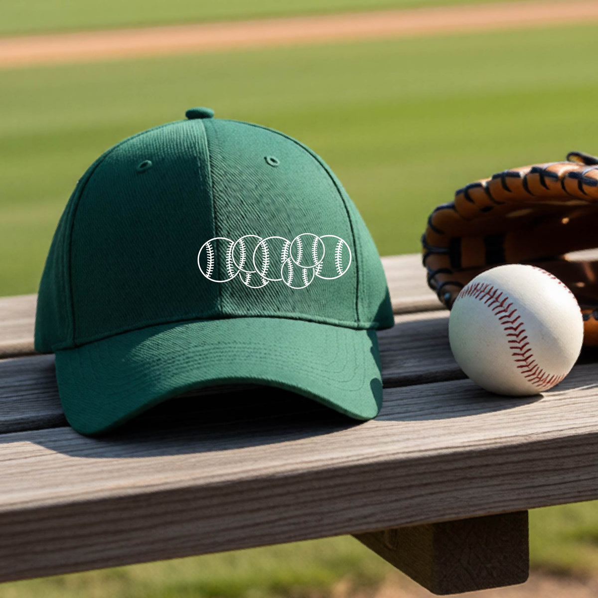 A closeup of a green baseball hat on an outdoor bench in front of a baseball diamond, with a white graphic of 6 sketched baseballs overlapping, printed on the front of the shirt