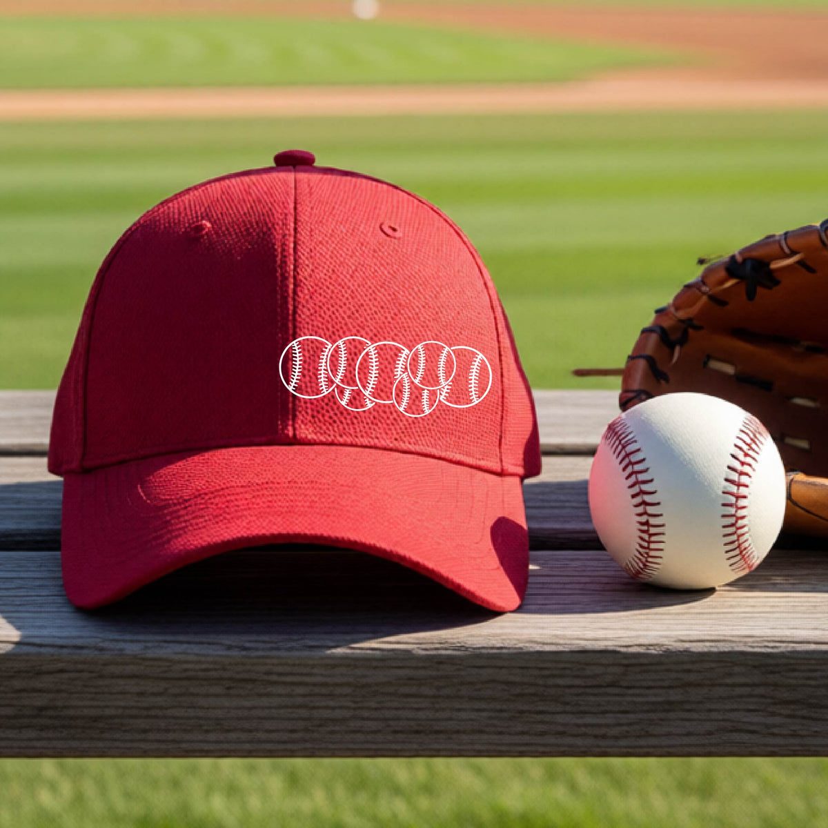 A closeup of a red baseball hat on an outdoor bench in front of a baseball diamond, with a white graphic of 6 sketched baseballs overlapping, printed on the front of the shirt