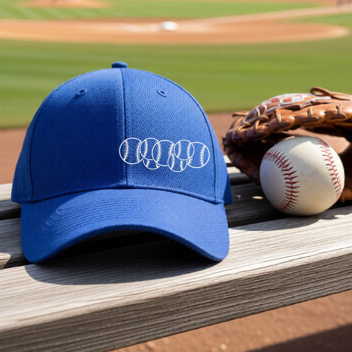 A closeup of a royal blue baseball hat on an outdoor bench in front of a baseball diamond, with a white graphic of 6 sketched baseballs overlapping, printed on the front of the shirt