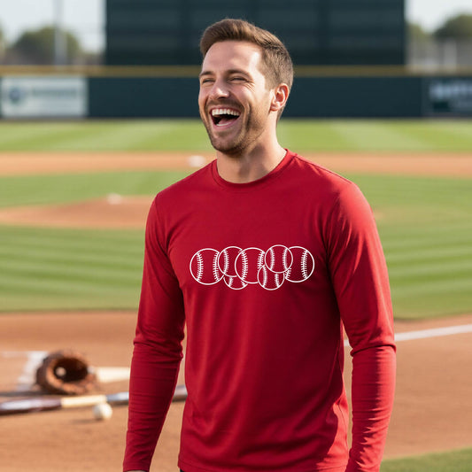 A man smiling in front of a baseball diamond wearing a red long-sleeve t-shirt, with a colorful graphic of a half baseball with a white graphic of 6 sketched baseballs overlapping, printed on the front of the shirt