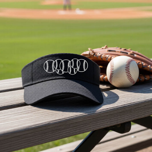 A close-up of a black baseball visor on an outdoor bench in front of a baseball diamond, with a white graphic of 6 sketched baseballs overlapping, printed on the front of the shirt