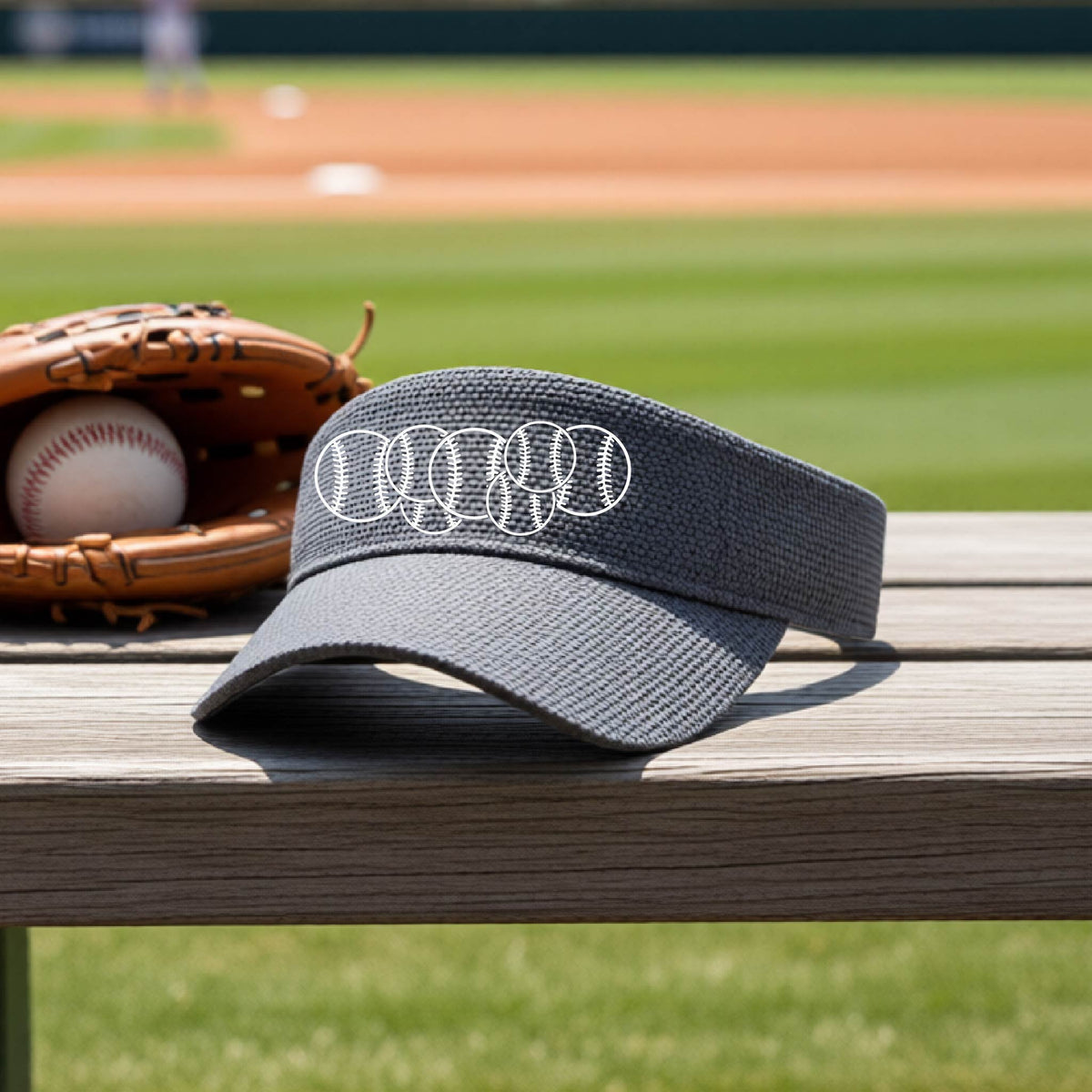 A close-up of a graphite gray baseball visor on an outdoor bench in front of a baseball diamond, with a white graphic of 6 sketched baseballs overlapping, printed on the front of the shirt