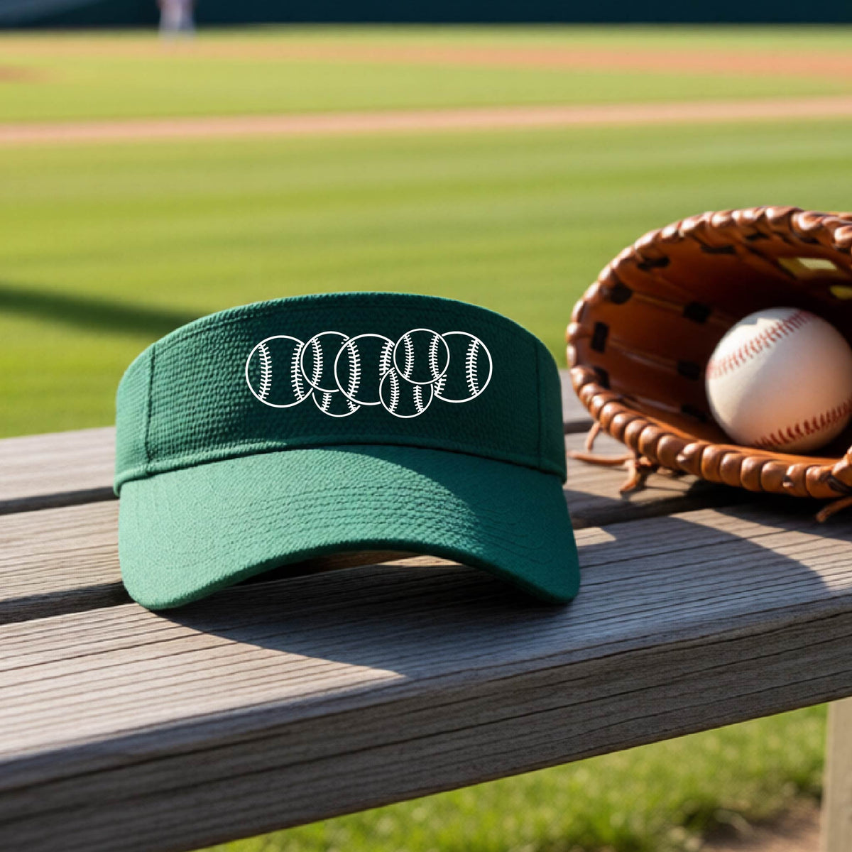 A close-up of a green baseball visor on an outdoor bench in front of a baseball diamond, with a white graphic of 6 sketched baseballs overlapping, printed on the front of the shirt