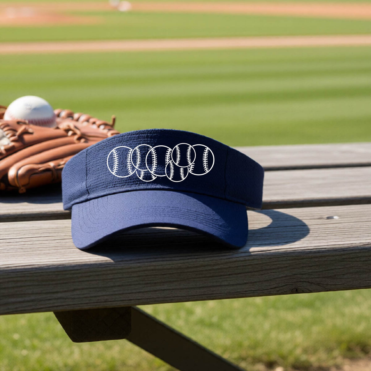 A close-up of a navy blue baseball visor on an outdoor bench in front of a baseball diamond, with a white graphic of 6 sketched baseballs overlapping, printed on the front of the shirt