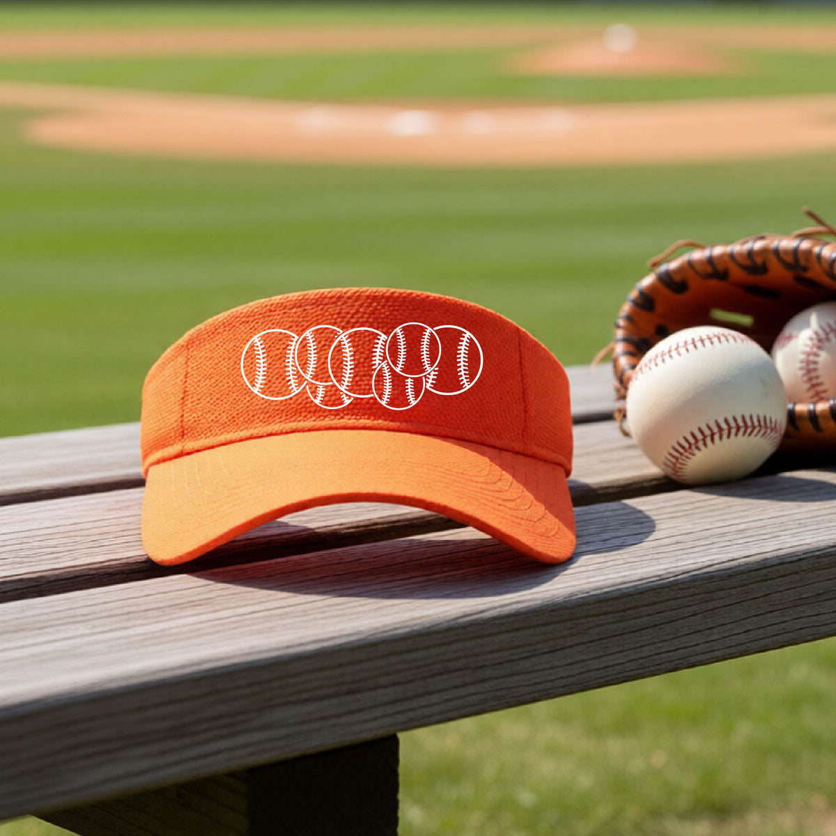 A close-up of an orange baseball visor on an outdoor bench in front of a baseball diamond, with a white graphic of 6 sketched baseballs overlapping, printed on the front of the shirt