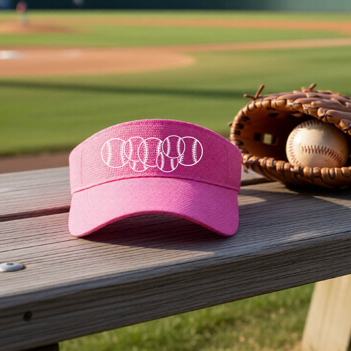 A close-up of a pink baseball visor on an outdoor bench in front of a baseball diamond, with a white graphic of 6 sketched baseballs overlapping, printed on the front of the shirt