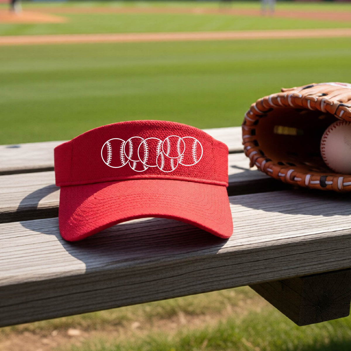 A close-up of a red baseball visor on an outdoor bench in front of a baseball diamond, with a white graphic of 6 sketched baseballs overlapping, printed on the front of the shirt