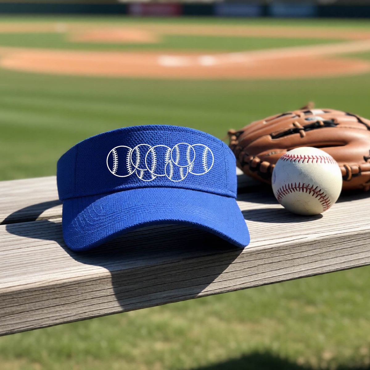 A close-up of a royal blue baseball visor on an outdoor bench in front of a baseball diamond, with a white graphic of 6 sketched baseballs overlapping, printed on the front of the shirt