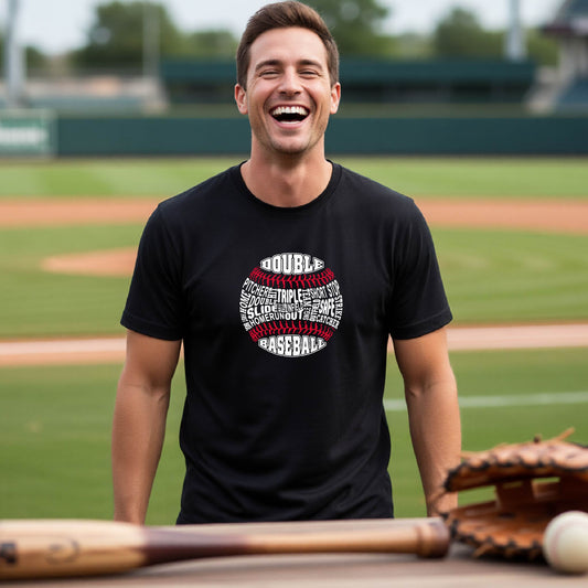 A man smiling in front of a baseball diamond wearing a black short-sleeve t-shirt, with a white and red graphic of a baseball made up of baseball words, printed on the front of the shirt.