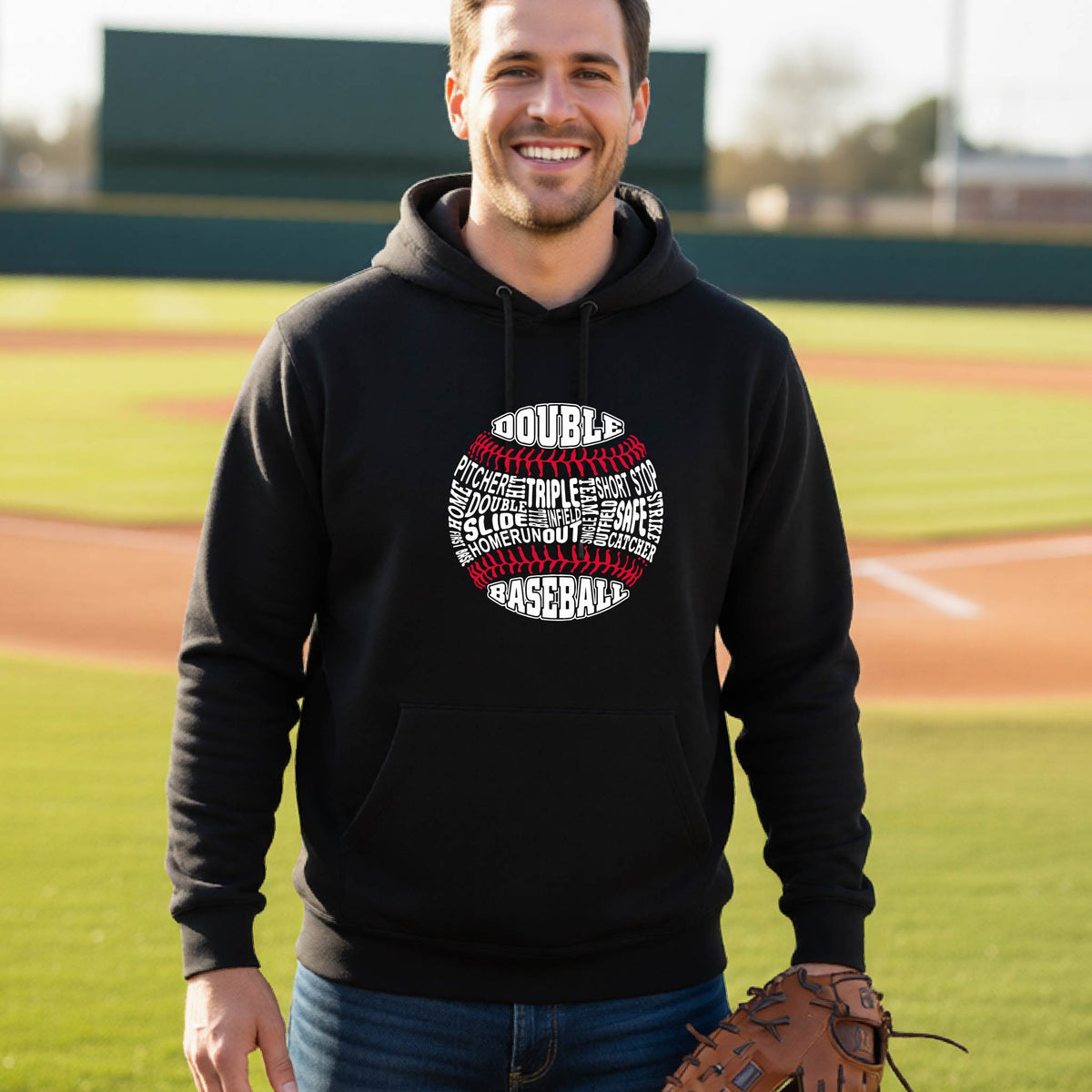A man smiling in front of a baseball diamond wearing a black pullover hoodie, with a white and red graphic of a baseball made up of baseball words, printed on the front of the shirt.