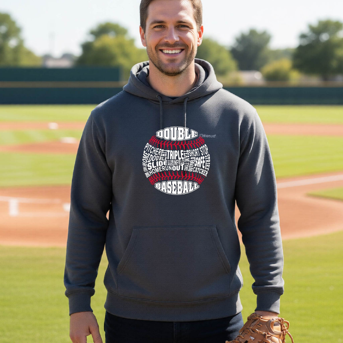 A man smiling in front of a baseball diamond wearing a gray pullover hoodie, with a white and red graphic of a baseball made up of baseball words, printed on the front of the shirt.
