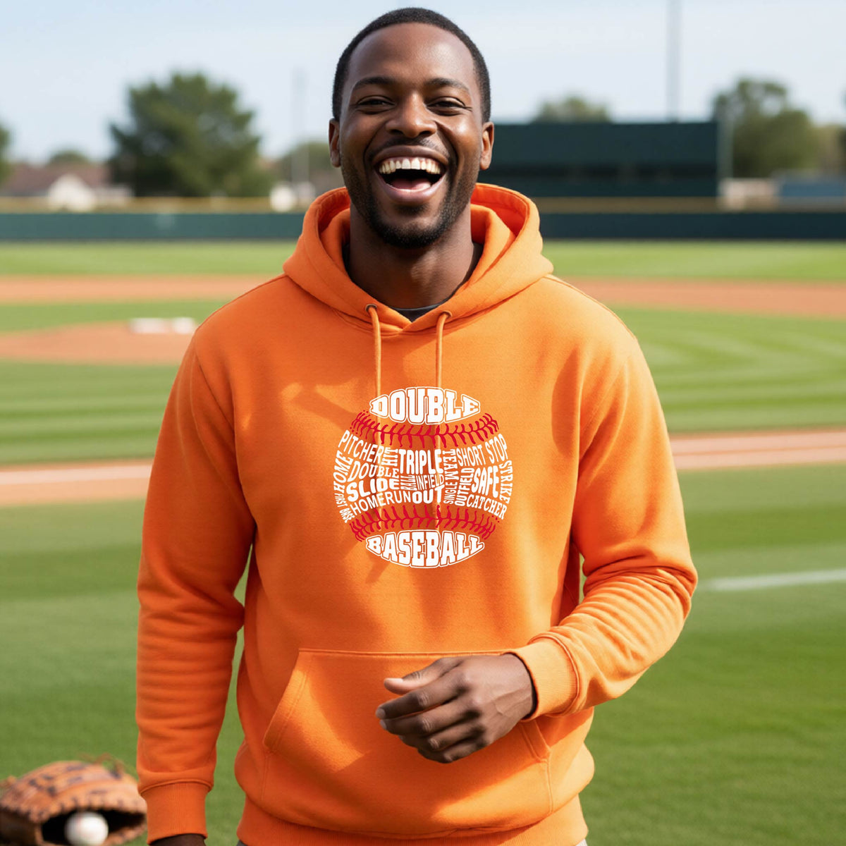 A man smiling in front of a baseball diamond wearing an orange pullover hoodie, with a white and red graphic of a baseball made up of baseball words, printed on the front of the shirt.