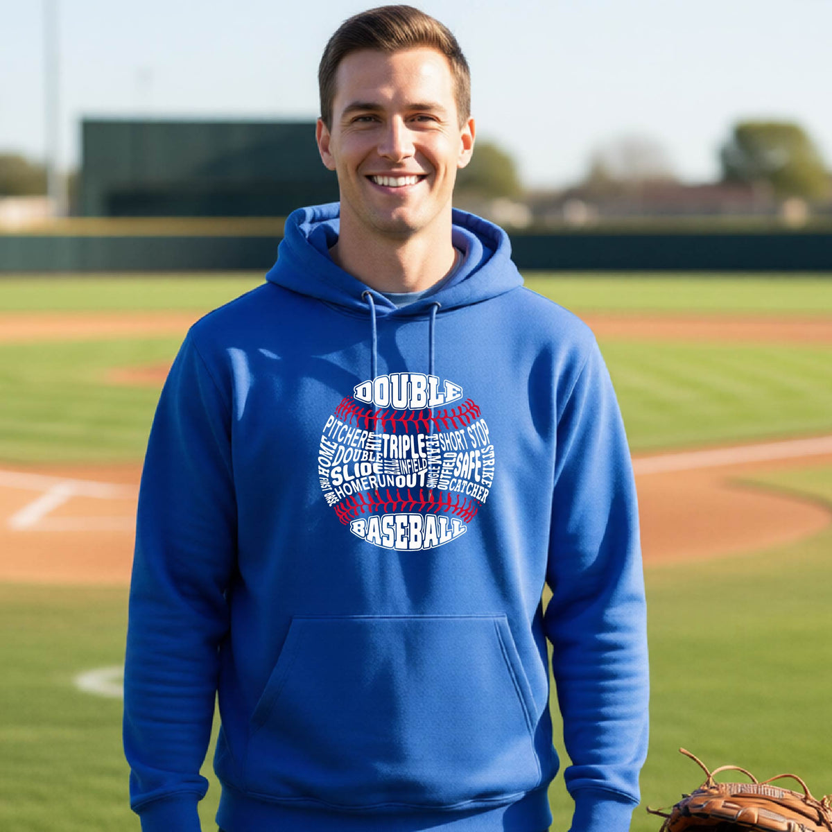 A man smiling in front of a baseball diamond wearing a royal blue pullover hoodie, with a white and red graphic of a baseball made up of baseball words, printed on the front of the shirt.