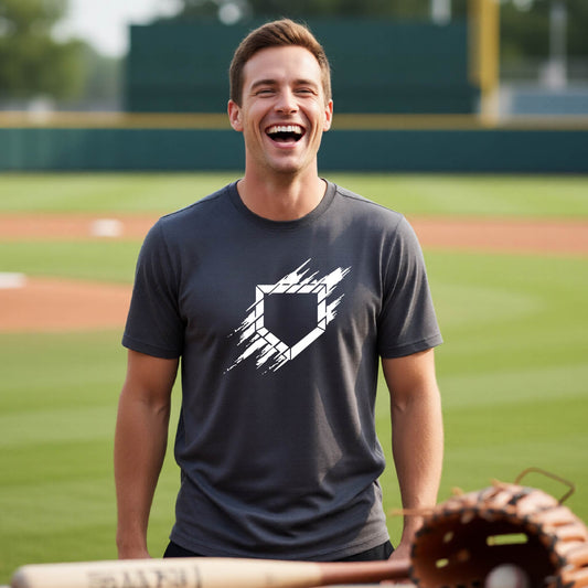 A man smiling in front of a baseball diamond wearing a dark gray short-sleeve t-shirt, with an artistic white graphic of a sketch of baseball home plate, printed on the front of the shirt.