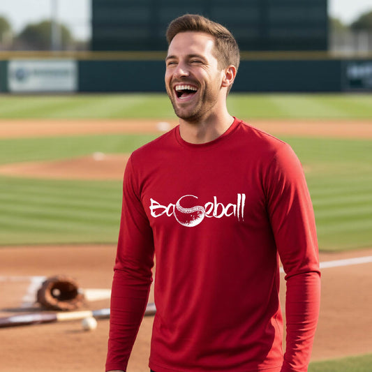 A man smiling in front of a baseball diamond wearing a red long-sleeve t-shirt, with an artistic white graphic of a sketch of the word BASEBALL” in a curved font with the S replaced by a baseball, printed on the front of the shirt.
