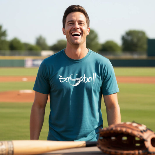 A man smiling in front of a baseball diamond wearing a teal short-sleeve t-shirt, with an artistic white graphic of a sketch of the word BASEBALL” in a curved font with the S replaced by a baseball, printed on the front of the shirt.